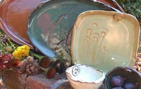 A collection of ceramic dishes and bowls in various earthy tones is displayed outdoors on a brick surface. Surrounding the ceramics are assorted dried flowers and fresh purple plums, giving the scene a rustic and natural feel.