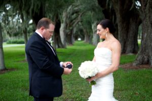 A bride and groom stand outdoors under a canopy of trees. The groom, in a black tuxedo, is looking at an item in his hands. The bride, in a white strapless gown, holds a bouquet of white flowers and looks at the groom with a smile.