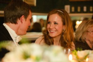 A woman with long, wavy brown hair and a man with short hair are sitting at a table, smiling and conversing. The table is adorned with white flowers, and another person is faintly visible in the background. The setting appears warm and festive.