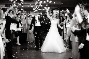 A black and white photo shows a bride and groom walking hand in hand down a path as wedding guests on either side cheer and throw flower petals. The bride is wearing a white dress and the groom a dark suit. The atmosphere is joyful and celebratory.