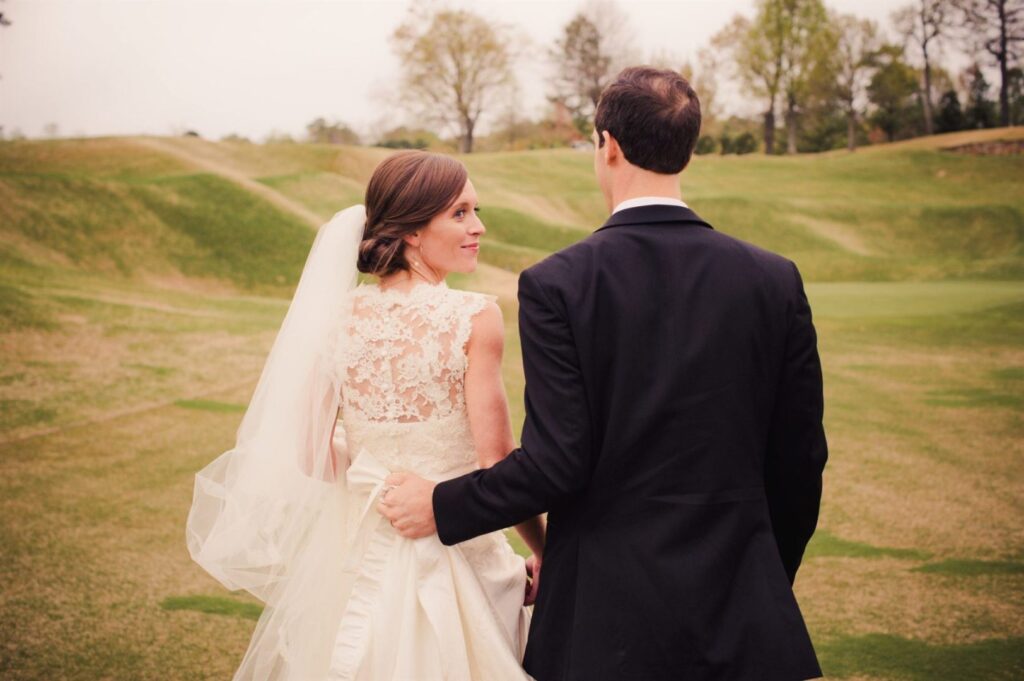 A bride and groom are standing together, with the bride in a white lace wedding dress and veil, and the groom in a black suit. They are facing away from the camera and holding hands, while the bride looks back over her shoulder, both surrounded by a scenic outdoor setting.