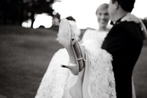 A grayscale image of a person in a suit carrying another person wearing a wedding dress. The focus is on the shoe of the individual in the wedding dress, showing its sole. The background appears to be an outdoor setting with trees and grass.