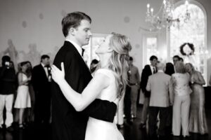 A couple dances closely at their wedding reception. The groom is in a black suit, and the bride is in a white gown. Guests are mingling in the background. The venue features a large, arched window adorned with a wreath and an elegant chandelier.