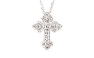 A close-up image of an ornate cross-shaped necklace pendant made of silver, featuring intricate floral patterns and multiple small embedded gemstones, suspended on a delicate chain.