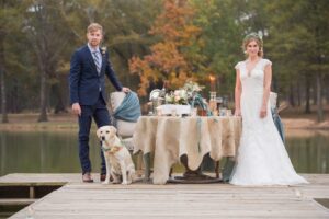 A couple stands on a wooden dock with a decorated table between them, overlooking a serene lake and surrounded by autumn foliage. The groom wears a navy suit while the bride is in a white wedding gown. A Labrador dog with a floral collar sits near the groom.