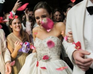 A joyful bride in a white strapless wedding gown smiles as pink rose petals are thrown in the air. She is surrounded by friends and guests celebrating with her, capturing a joyous moment of the wedding reception.