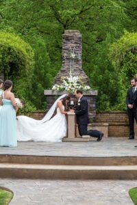 A bride and groom kneel in front of an outdoor altar during their wedding ceremony. The bride wears a white dress and veil, while the groom is in a black suit. Bridesmaids in light blue dresses and a groomsman in black are standing nearby. Greenery surrounds the altar.