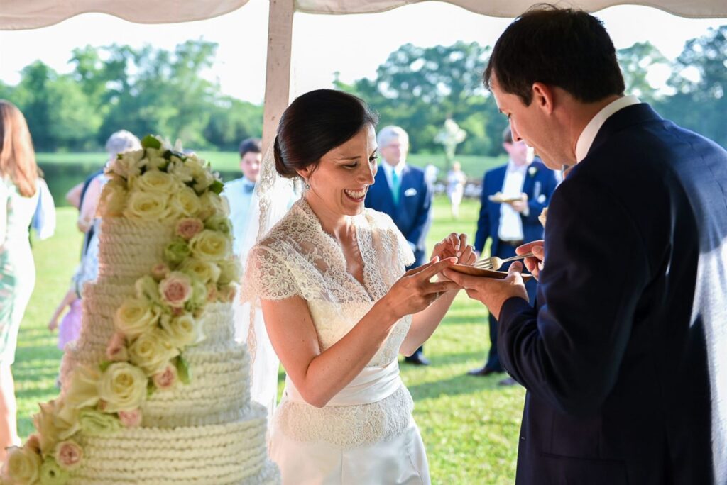 A bride and groom are sharing a moment during their wedding reception under a tent. The bride, dressed in a lace gown, smiles as she holds a piece of cake while the groom, in a suit, prepares to take a bite. A tiered wedding cake adorned with flowers is visible.