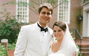 A smiling bride and groom stand close together outdoors on their wedding day. The groom is dressed in a white tuxedo with a black bow tie, and the bride is wearing a white wedding dress and veil, holding a bouquet of white flowers.