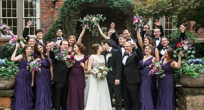 A wedding party gathers in front of a brick building adorned with greenery. The bride and groom stand in the center, surrounded by bridesmaids in purple dresses and groomsmen in black suits, all cheering and holding bouquets aloft.