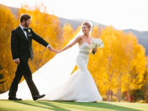 A bride in a white wedding dress holds a bouquet of white flowers and walks hand in hand with a groom in a black tuxedo. They are smiling at each other, with a backdrop of vibrant yellow autumn trees and mountains under a clear sky.