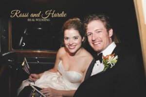 A happy couple, dressed in wedding attire, sits closely together inside a vehicle. The bride wears a strapless white dress and holds a small American flag, while the groom, in a tuxedo with a boutonniere, smiles brightly. Text reads: "Rossi and Hardie, Real Wedding.