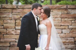 A groom in a black tuxedo gently kisses a bride's forehead as she closes her eyes. The bride, in a white strapless gown and veil, holds a bouquet of white flowers. They stand in front of a stone wall, sharing an intimate moment on their wedding day.