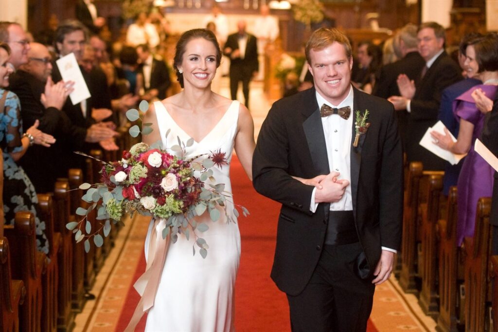 A bride in a white gown and a groom in a black tuxedo walk arm-in-arm down the aisle of a church, smiling. The bride holds a bouquet of flowers, and guests on both sides of the aisle stand and clap. The church is warmly lit, with a red carpet and wooden pews.