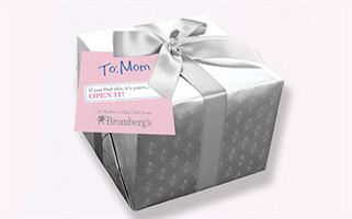 A square gift box wrapped in elegant silver paper and topped with a silver ribbon bow. Attached is a pink and white tag reading "To: Mom" and an additional message suggesting to open if specific conditions are met. The tag is branded with Bromberg's logo.