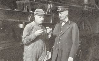 A black-and-white photo shows two men standing and talking in front of a large piece of machinery. The man on the left is dressed in work overalls and a cap, while the man on the right is wearing a conductor's uniform and hat, holding a pocket watch.