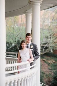 A couple dressed in wedding attire stands on a white, curved balcony. The bride is wearing a white lace dress, and the groom is in a dark suit with a floral boutonniere. They are smiling, and trees with new leaves are visible in the background.