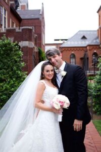 A bride and groom stand close together, smiling for a wedding photo in an outdoor area. The bride is wearing a white wedding dress and holding a bouquet of pink and white flowers. The groom is in a black suit and bow tie. There are brick buildings in the background.