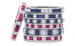 A stack of seven rings is shown, featuring alternating rows of brightly colored gemstones. The gemstones include red, blue, and clear stones, each set in a silver band. The rings are intricately designed and neatly aligned.