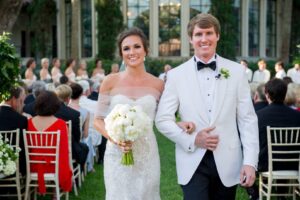 A bride in a strapless wedding gown holds white flowers, and a groom in a white tuxedo with a black bow tie, smile while walking down the aisle outdoors. Guests seated behind them watch the couple. Bridesmaids in light-colored dresses stand in the background.