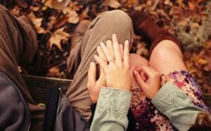 Two people are sitting closely on a bench, their hands tenderly entwined. One person wears a green jacket, the other a floral dress. Fallen autumn leaves surround them, adding a warm, rustic background.