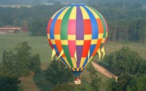 A colorful hot air balloon with a checkered pattern of blue, yellow, green, red, and pink is floating above a green landscape with trees and a dirt path below. The basket of the balloon is visible, carrying passengers.