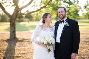 A bride and groom smile at each other while standing outdoors on a sunny day. The bride is wearing a white lace wedding dress and holding a bouquet of white flowers. The groom is dressed in a black tuxedo with a bow tie and a boutonniere. Trees are in the background.