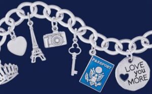 A silver charm bracelet features various charms including a heart, Eiffel Tower, camera, key, passport, crown, and a round pendant inscribed with "LOVE YOU MORE" and a heart cutout. The background is solid dark blue.