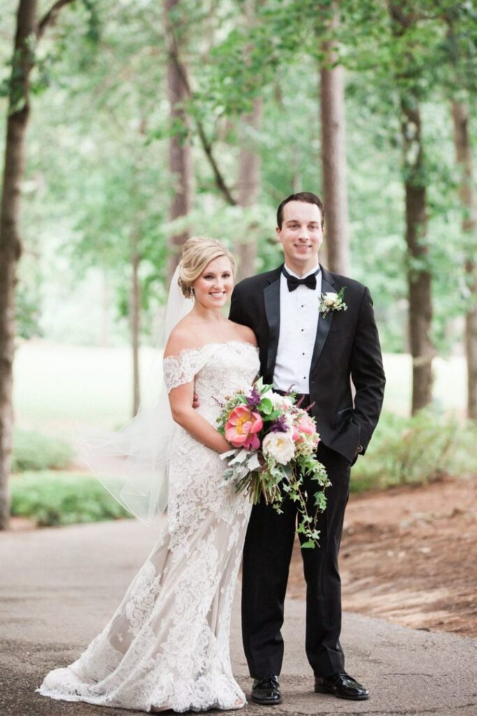 A bride and groom stand together outdoors for a wedding photo. The bride is wearing an off-the-shoulder lace dress and holding a bouquet of colorful flowers. The groom is dressed in a black tuxedo with a bow tie. They are surrounded by trees and greenery.