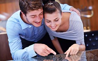 A smiling man and woman are leaning closely together, both pointing and looking at something on a glass surface in front of them. The woman has her hair tied back, and they appear to be engaged and happy.