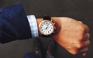 A person wearing a plaid shirt and blue jacket glances at a watch on their left wrist. The watch has a white face with black numerals and a brown leather strap. The person's hand is adorned with a ring, and the ground below is gray and slightly textured.