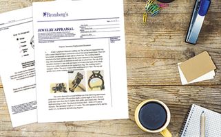 Close-up view of a wooden desk with scattered items including a jewelry appraisal document with images of jewelry, a cup of coffee, a spiral notebook, a stapler, colorful paper clips, and several papers and business cards.