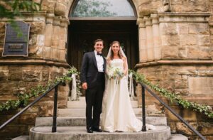 A bride and groom stand on the steps in front of a church. The groom is wearing a black tuxedo, and the bride is in a white wedding dress holding a bouquet of flowers. They are both smiling and standing under an archway decorated with greenery.