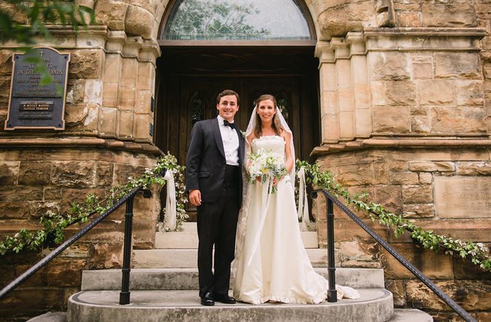 A bride and groom stand on the steps in front of a church. The groom is wearing a black tuxedo, and the bride is in a white wedding dress holding a bouquet of flowers. They are both smiling and standing under an archway decorated with greenery.