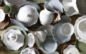 A scattered assortment of ceramic dishes, including plates, bowls, cups, and pitchers—a perfect addition to any wedding registry. The dishes come in various shades of white, light blue, green, and brown with both plain and scalloped edges. This artistic display sits elegantly on a textured surface.