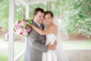 A bride in a white wedding dress and veil holds a bouquet of pink and white flowers while hugging a groom in a gray suit and striped tie. They are standing on a white porch with green trees in the background, smiling at the camera.