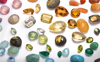 A collection of various gemstones in different shapes, sizes, and colors spread out on a white surface. The stones include shades of green, blue, orange, red, yellow, and clear, with cuts such as oval, round, pear, and rectangular, showcasing vibrant hues and facets.