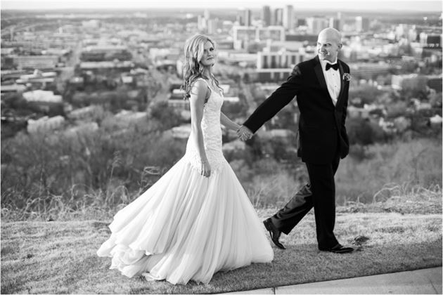 A black and white photo of a newlywed couple holding hands and walking along a scenic overlook. The bride wears a flowing gown, and the groom is in a tuxedo. City buildings and trees are visible in the background, with the couple smiling at each other.