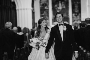 A bride and groom walk down the aisle holding hands in a church. The bride wears a white dress and a tiara, holding a bouquet of flowers, while the groom is in a tuxedo with a boutonniere. Both are smiling, and there are guests seated on either side of the aisle.