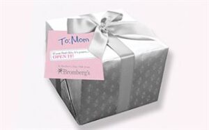 A silver and white-wrapped gift box with a silver ribbon and bow. Attached is a pink and white tag addressed "To: Mom" with additional text instructing to open the gift. The packaging has a subtle floral pattern and the tag includes the logo for Bromberg's.