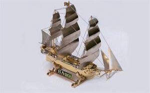 A detailed, miniature metal ship model with numerous sails and intricate rigging. The metallic finish of the ship shines, highlighting its finely crafted structure. The base of the ship features a plaque with the inscription "S.S. ZERO.