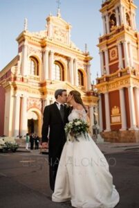 A bride and groom stand in front of a historic church with pink and gold detailing. The bride is in a white gown holding a bouquet, and the groom is in a black suit. They are about to kiss, and a bell tower is visible in the background.