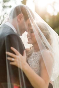 A bride and groom share an intimate moment under a veil on their wedding day. The bride is wearing an embellished gown, and the groom is in a black suit. They are embracing, with soft sunlight shining through the trees behind them, creating a warm and romantic atmosphere.