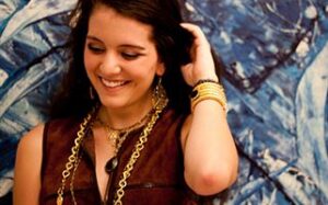 A woman with long dark hair smiles while looking down. She is wearing a brown sleeveless top and several gold necklaces and bracelets. She is standing in front of a blue and white abstract background.