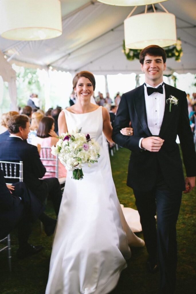 A smiling bride in a white gown and a groom in a black tuxedo walk arm-in-arm down the aisle in an outdoor tent. The bride holds a bouquet of white flowers and the groom has a boutonnière. Guests are seated and watching them in the background.