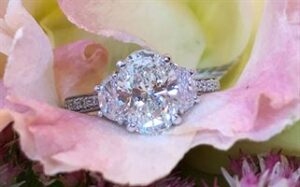 A sparkling diamond engagement ring with a central round-cut diamond flanked by two smaller diamonds is nestled within the delicate petals of a pink and yellow rose. The ring's band is encrusted with smaller diamonds, adding extra shimmer to the elegant setting.