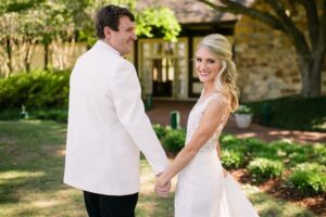 A couple dressed in wedding attire holds hands outdoors. The woman, in a white lace bridal gown, faces the camera and smiles, while the man, in a white suit jacket and black pants, looks at her. They are in a lush garden with trees and a stone building in the background.