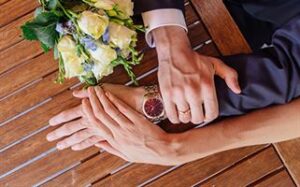 A close-up of a couple's hands on a wooden table. The person on the left wears a watch and a suit sleeve, while the person on the right has a hand resting on the other's arm. A bouquet of white and purple flowers is placed near their hands.