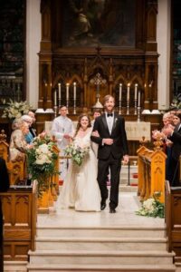 A bride and groom walk down the aisle of a church after getting married. The bride wears a white gown and holds a bouquet, while the groom is in a black tuxedo. Guests on both sides of the aisle stand and clap, and the altar is decorated with flowers and candles.