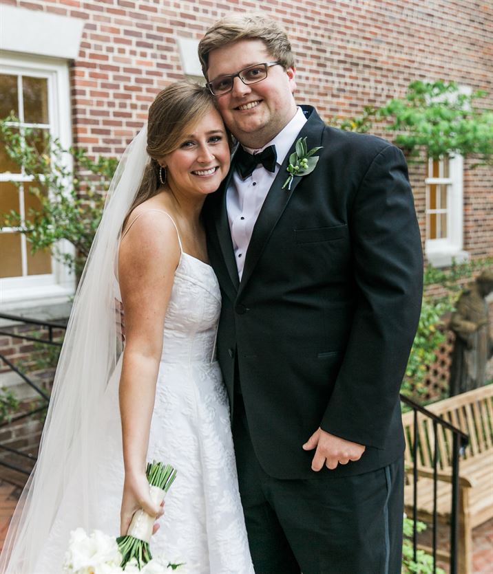 A bride and groom stand closely together in front of a brick building. The bride is wearing a white wedding gown and veil, holding a bouquet of flowers. The groom is dressed in a black tuxedo with a bow tie and boutonnière. They are both smiling happily.